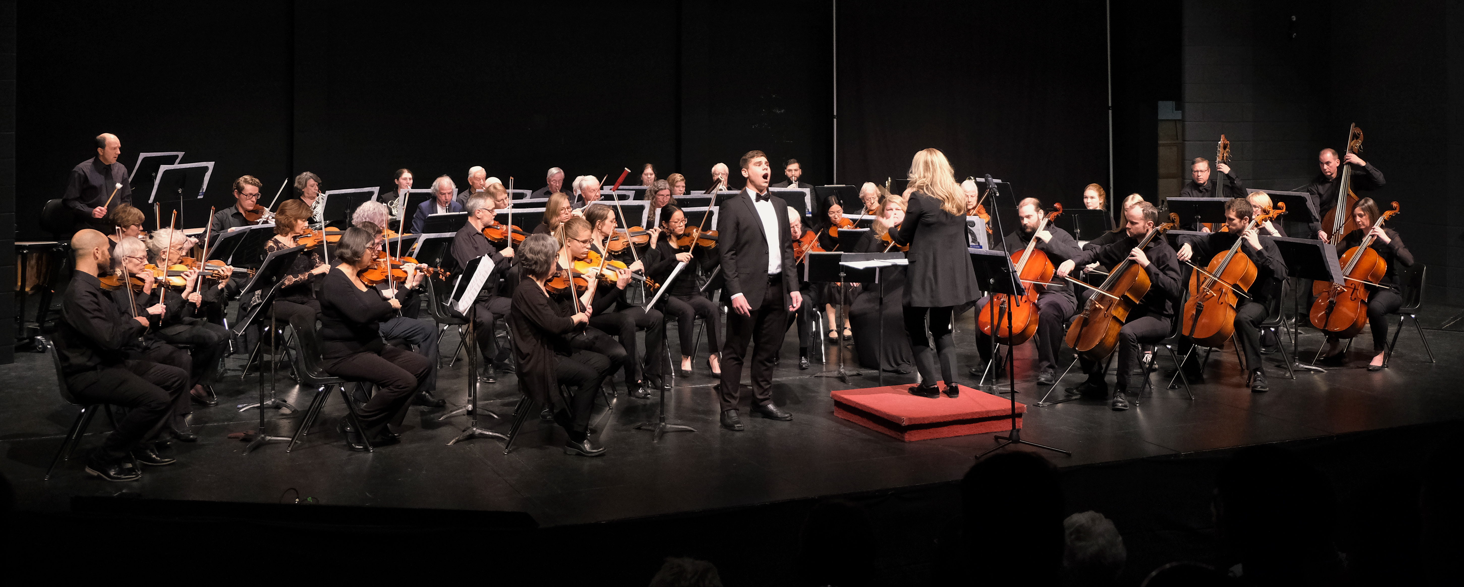Mennonite Community Orchestra in the middle of a performance with baritone soloist. Orchestra members are wearing black and arranged on a black stage.