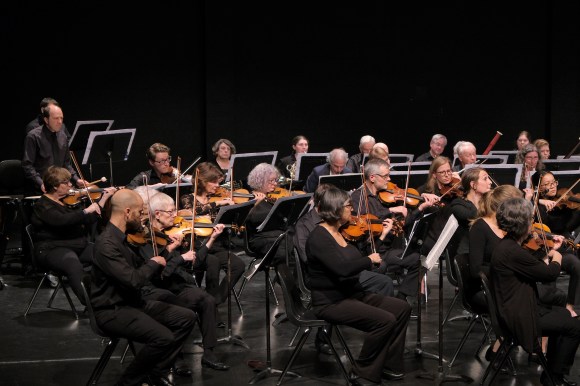 First and second violins playing during a concert at Jubilee place. Players are wearing black and sitting on a black stage.