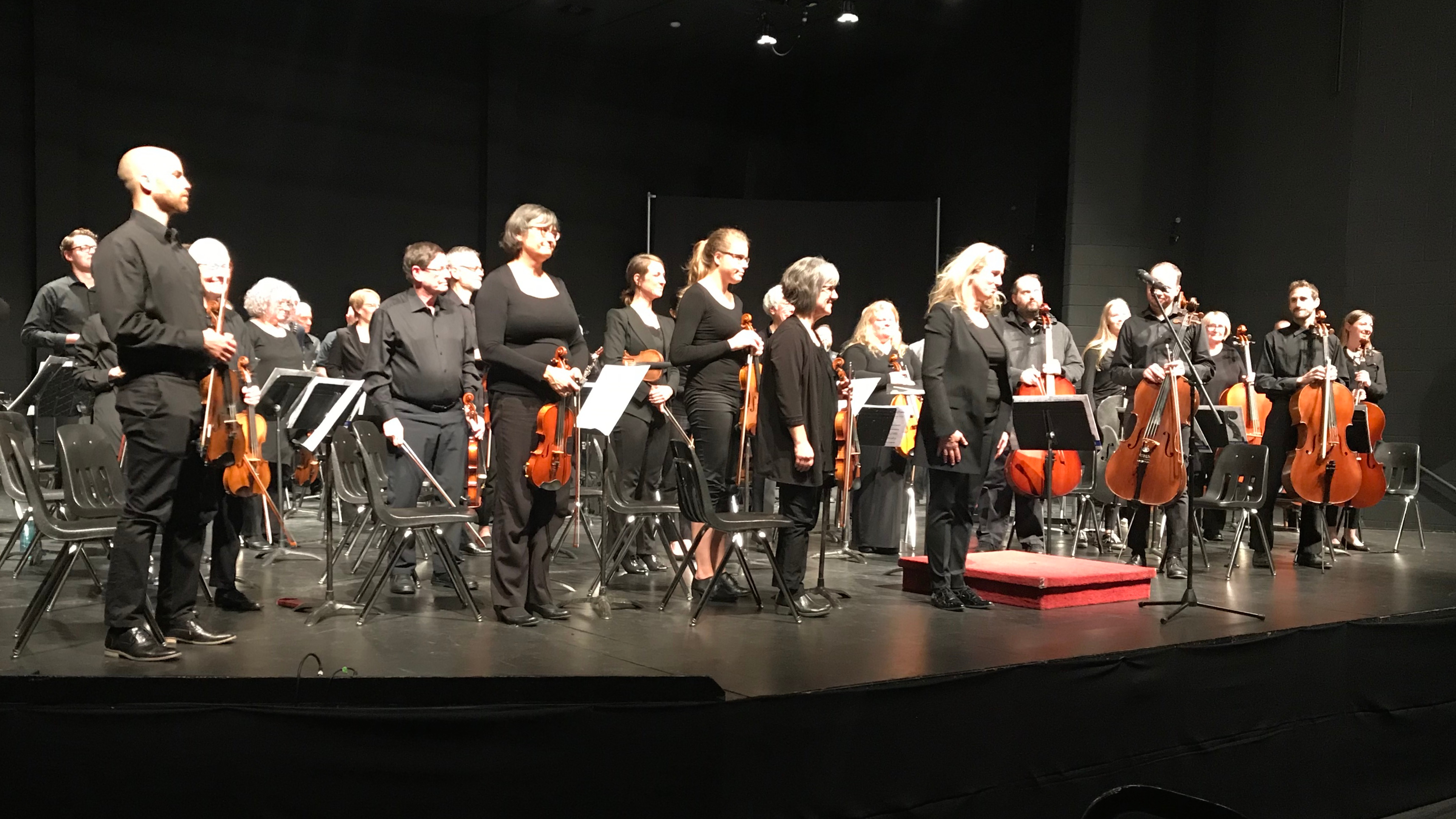 MCO players and conductor standing on a black stage (Jubilee Place) facing the audience.